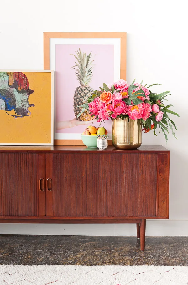 A wood sideboard with ridged sliding fronts and small oval pulls stands beneath leaning colorful artworks beside a gold-colored flower vessel