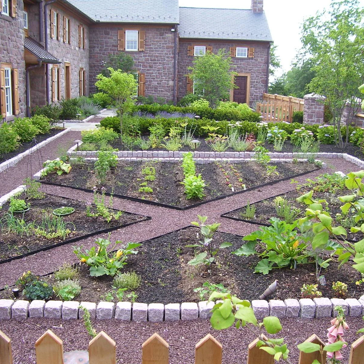 Low-edged vegetable beds divided by diagonal gravel paths beside a stone house and picket fence.