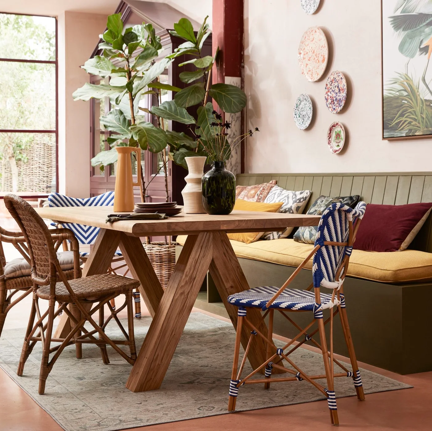 A green-backed cushioned dining bench sits beside pale warm walls and a wooden table with woven and blue-striped chairs