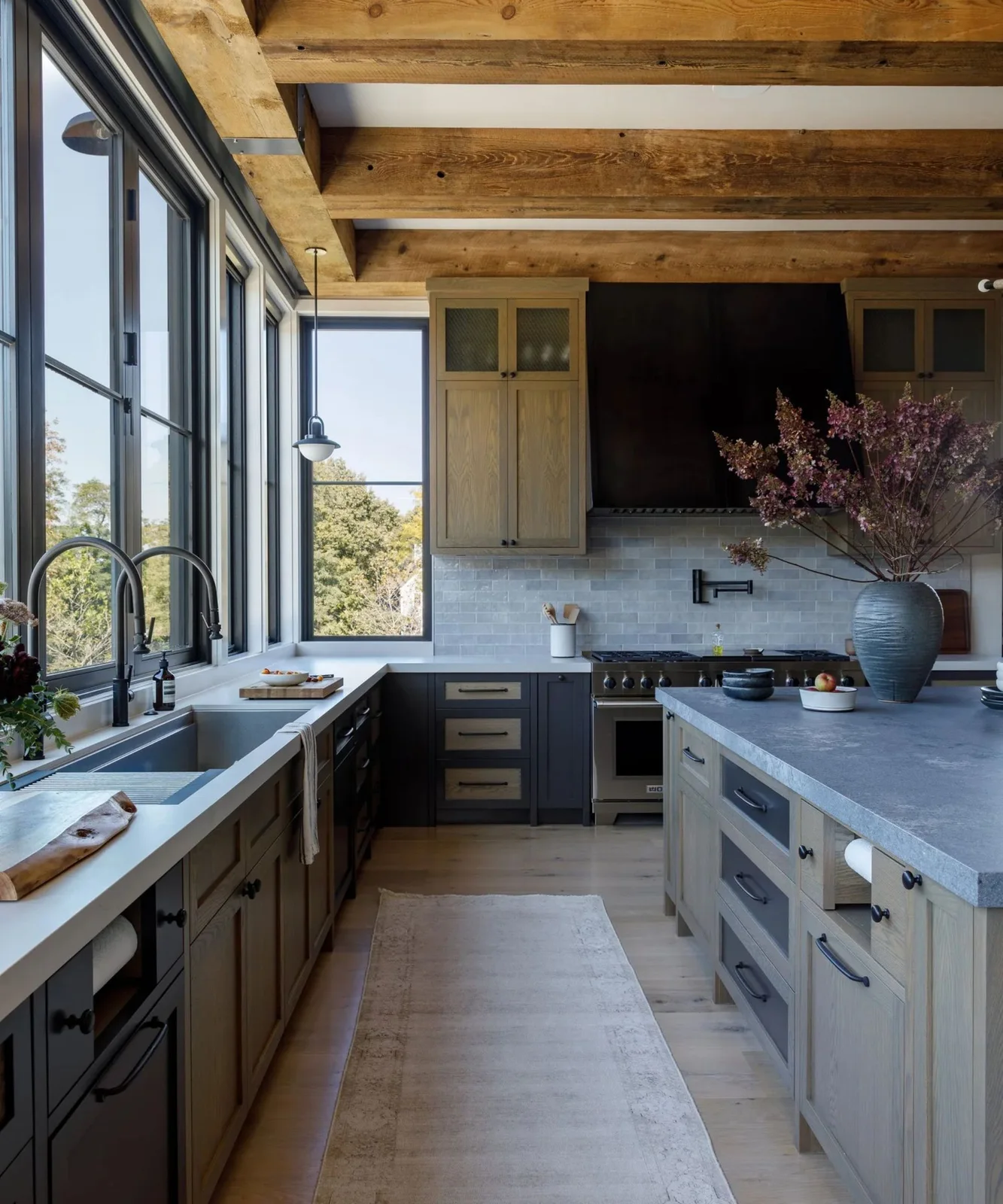 Natural oak cabinets mix with dark painted drawers beneath heavy wood beams, with a black hood, tall windows and pale counters.