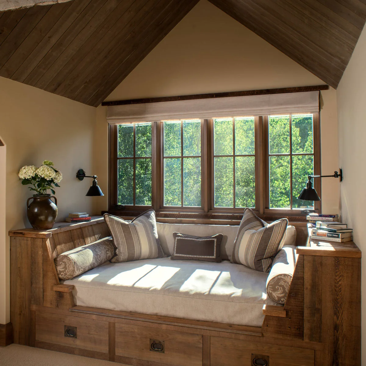 A wood built-in daybed has broad side ledges, lower drawers and pale cushions beneath a wide window.