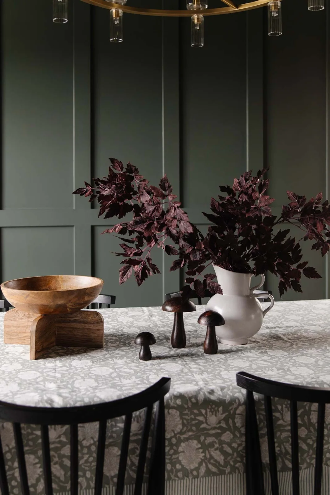 A pale patterned dining cloth holds burgundy branches in white pitchers, a wooden bowl and small mushroom-shaped ornaments against green paneling