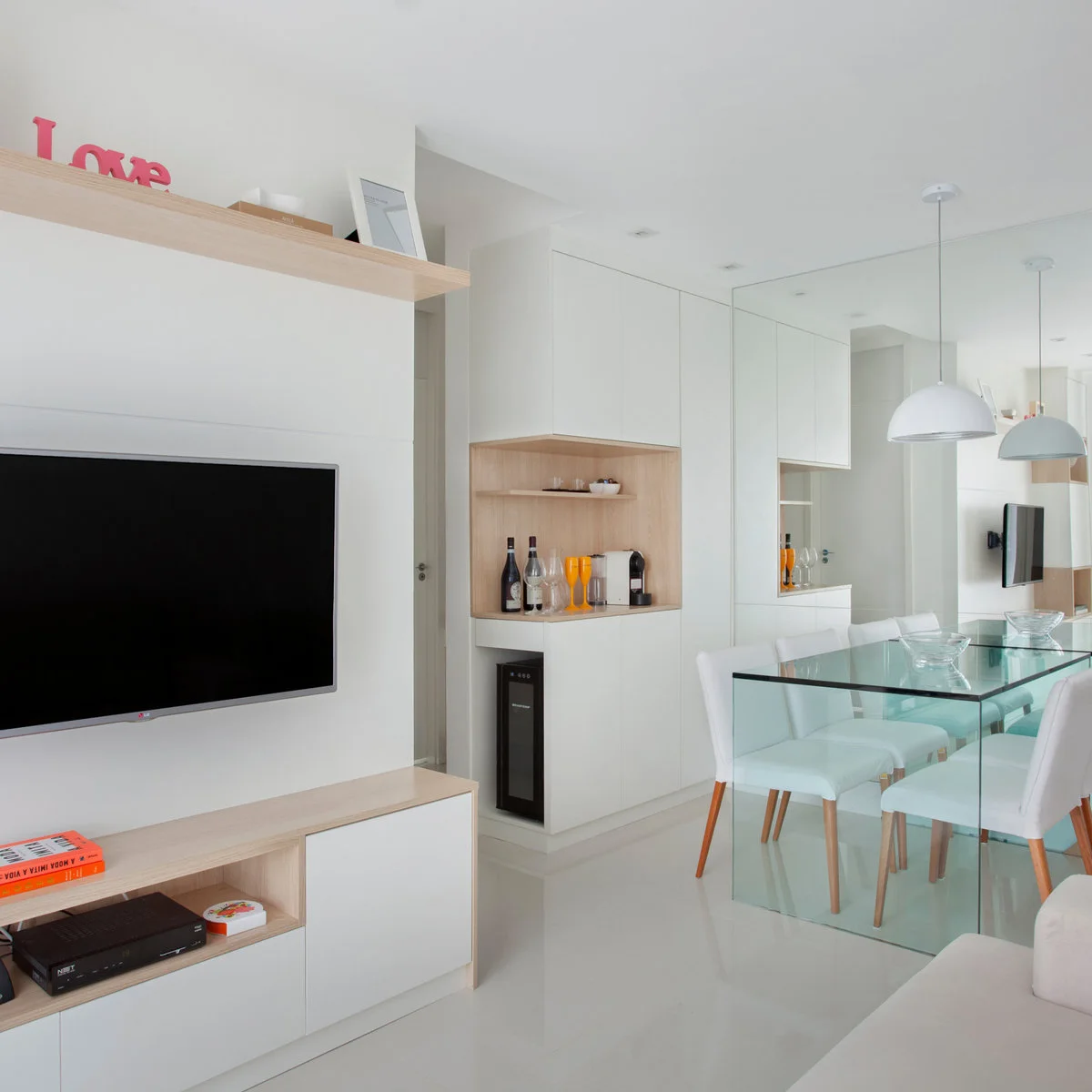 A compact pale room has a wall television above low storage, a glass dining table with white chairs and a large mirror behind the table.
