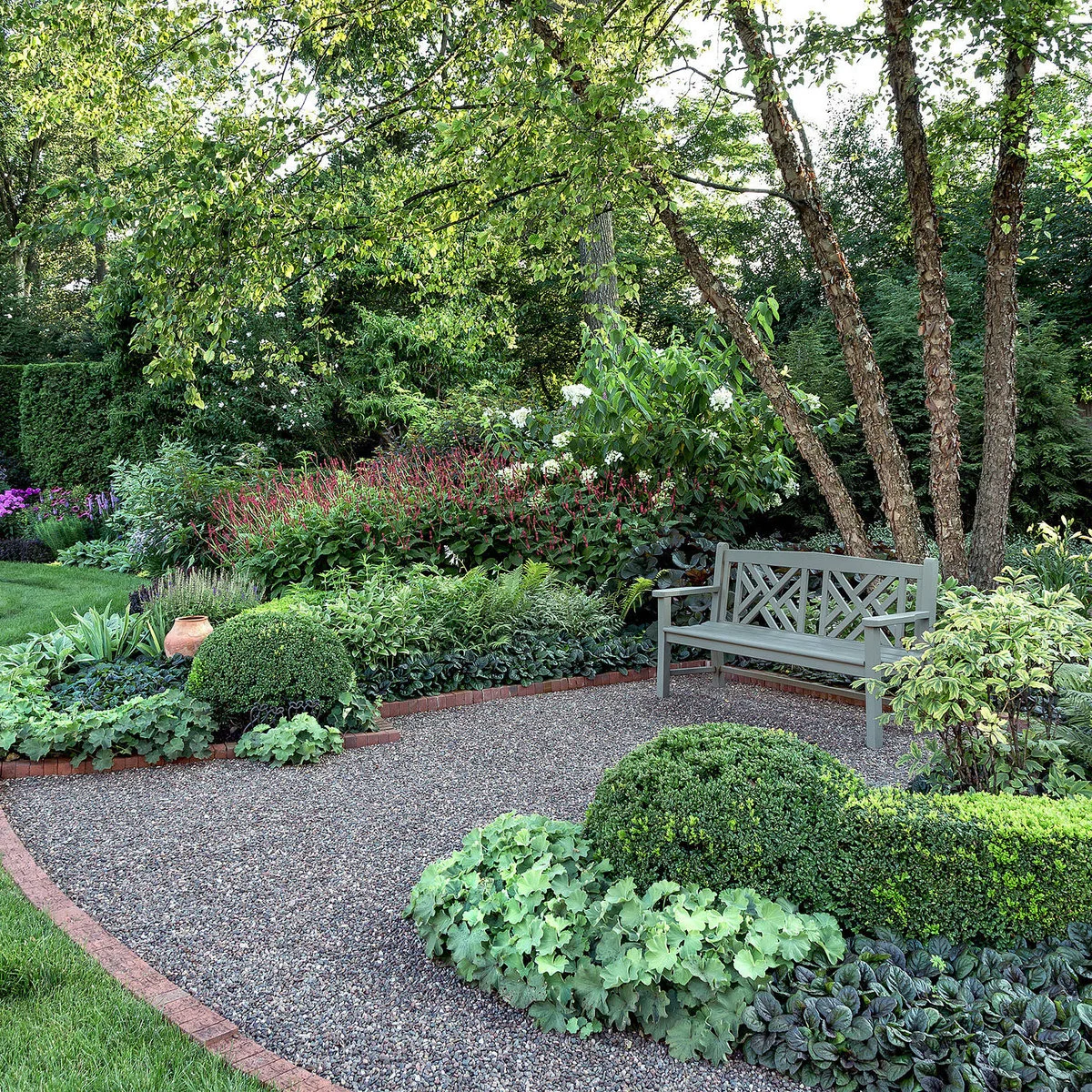 A gray garden bench stands on a curved gravel area beneath trees, surrounded by layered planting and low brick edging.