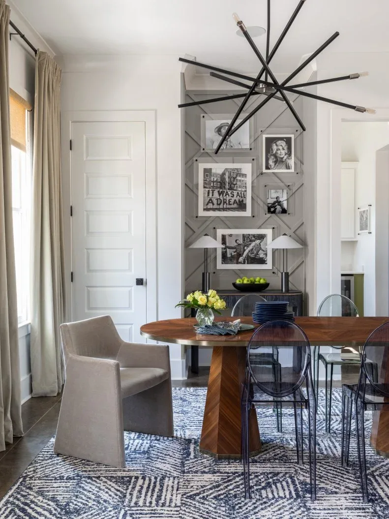 Gray narrow dining-room feature wall with diagonal trim, framed black-and-white pictures, low console and lamps, wood table, transparent chairs, and black chandelier.