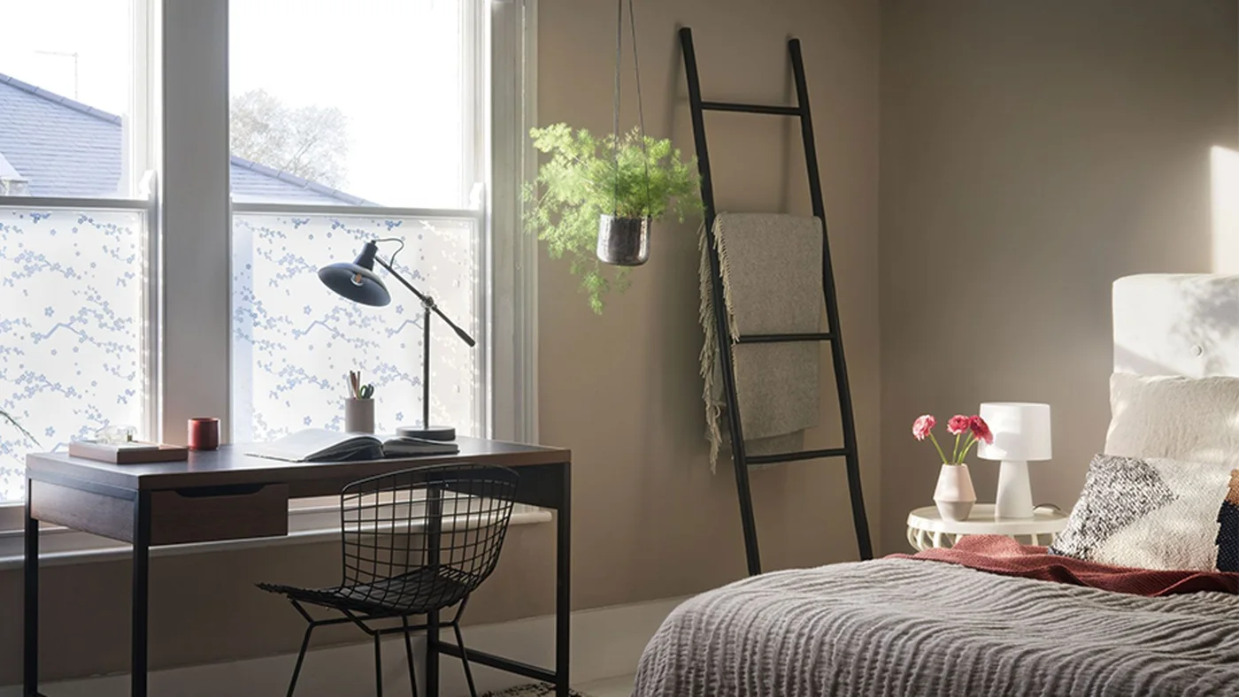 A dark desk with a wire chair sits beside a pale bed beneath tall windows, with a small task lamp and a dark textile ladder