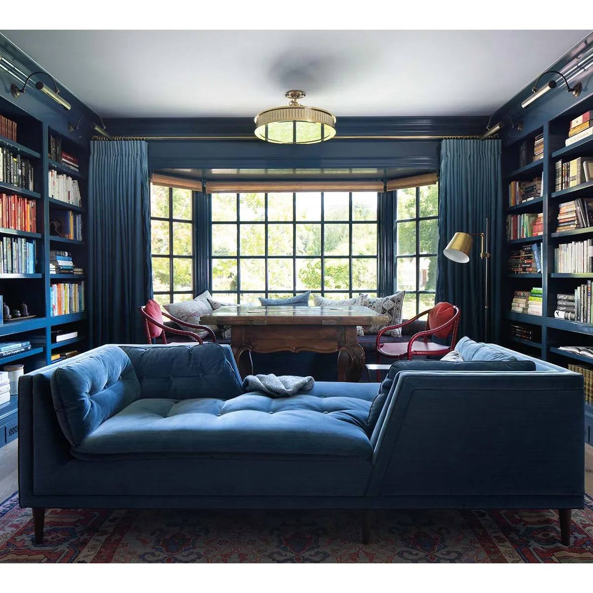 A deep blue tufted daybed with raised ends stands between blue bookcases and a desk by a bay window.