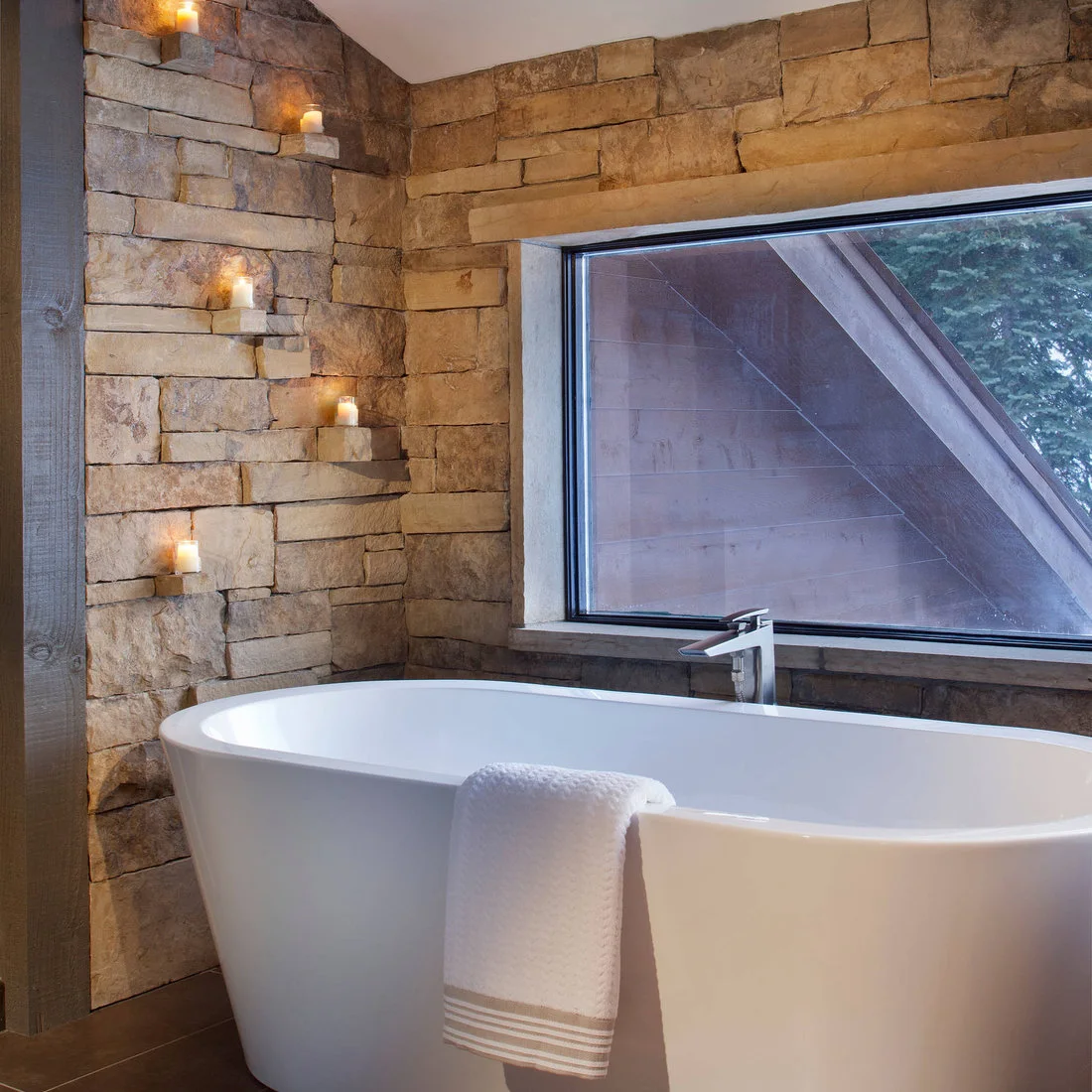 A white freestanding bath beside rough stone walls and a broad dark framed window