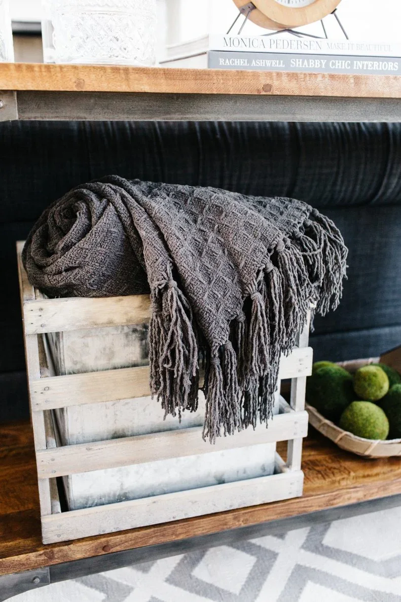 A pale slatted crate on a console shelf holds a rolled gray fringed throw against dark upholstery.