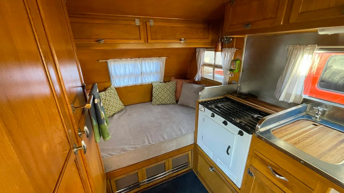 Vintage camper with honey-colored cabinetry, a gray cushioned end bench and a compact white cooker beside the sink.