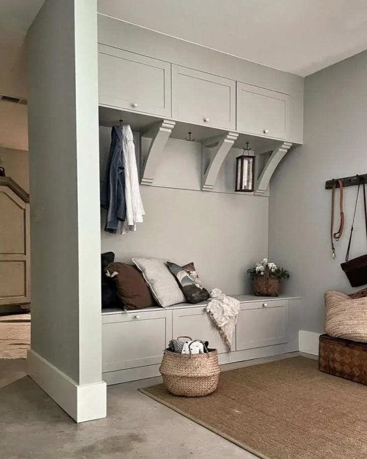 Soft gray built-in mudroom arrangement with upper cabinet doors, decorative supports, low storage and a cushioned sitting area.