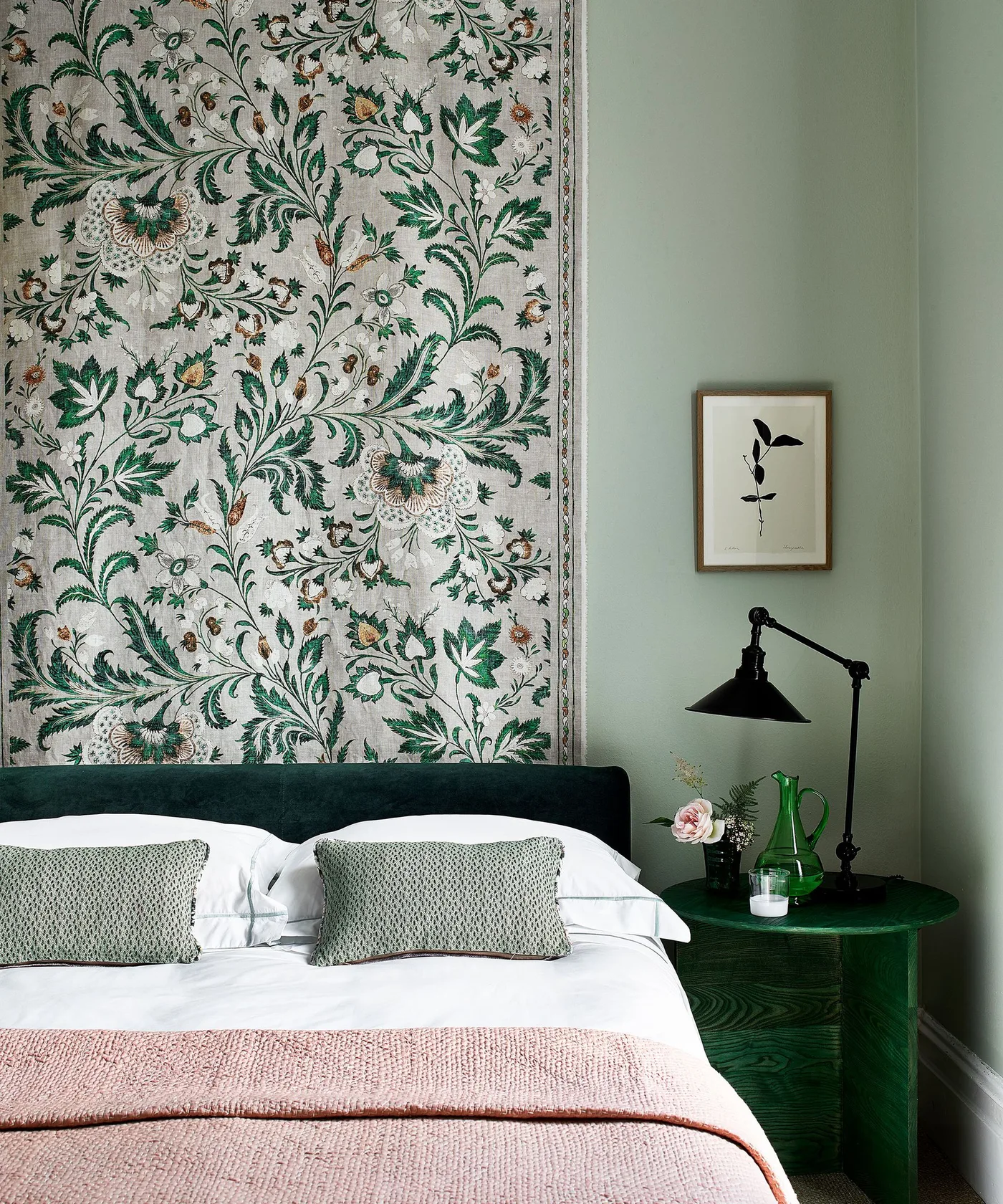 Bedroom with green floral wall hanging, dark green headboard and round bedside table, pale bedding, pink throw and adjustable dark lamp.