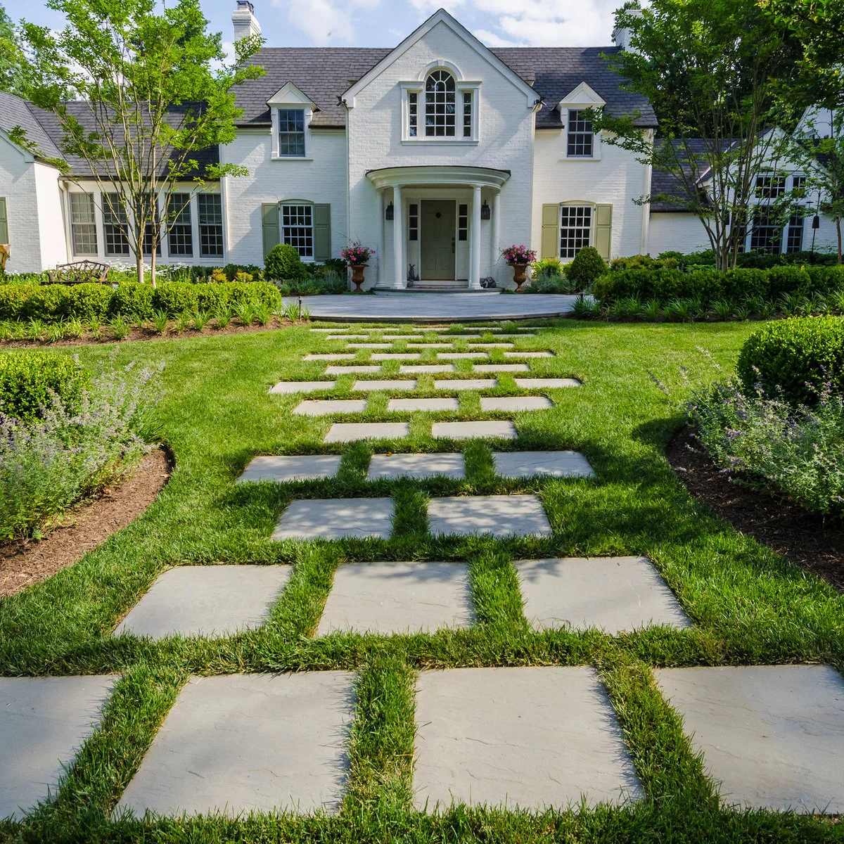 Large rectangular pavers set into grass lead toward the central portico of a white house with planting beds on both sides.