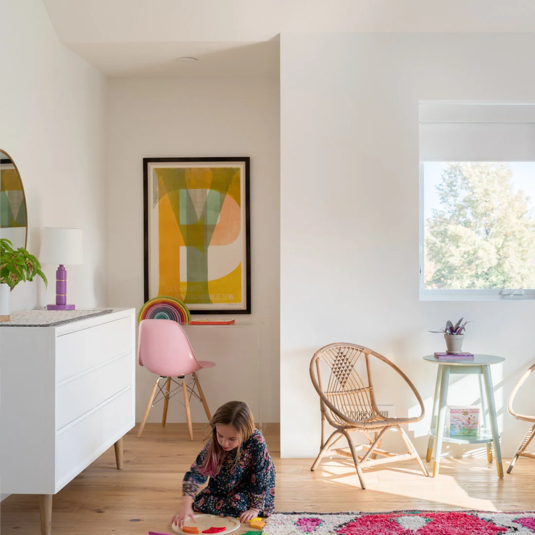 A child plays on a wood floor beside a white dresser, a small round table with woven chairs, a pink desk chair and a colorful rug.