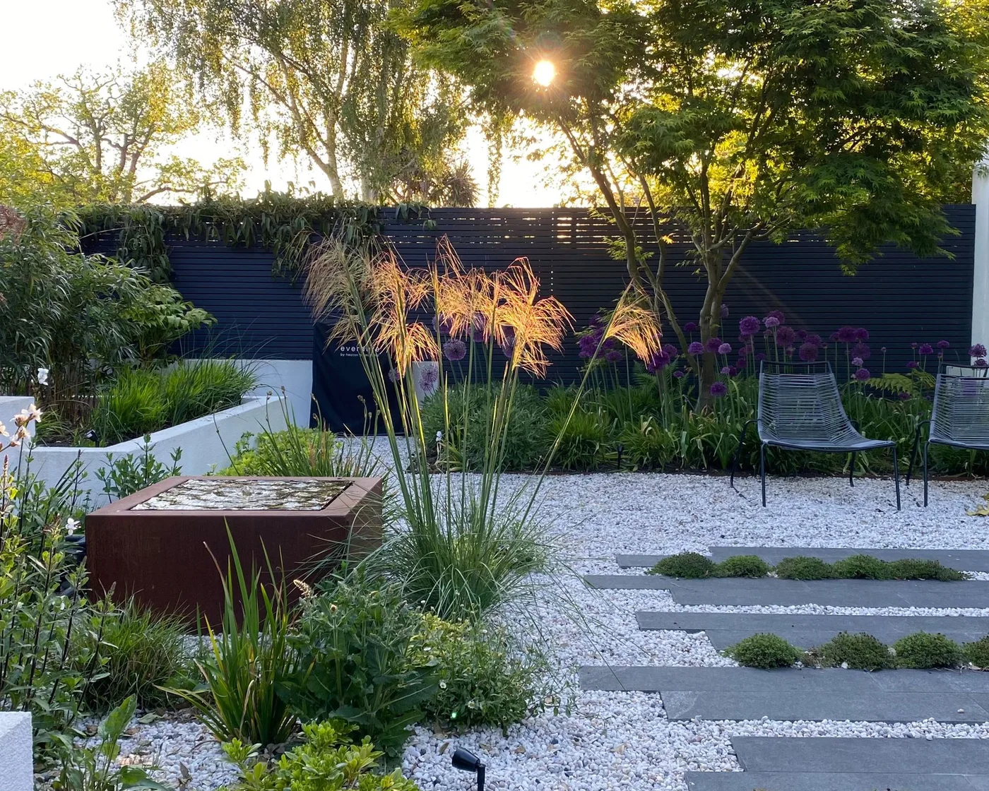 Dark stepping slabs cross pale gravel beside planted beds and a square raised water feature