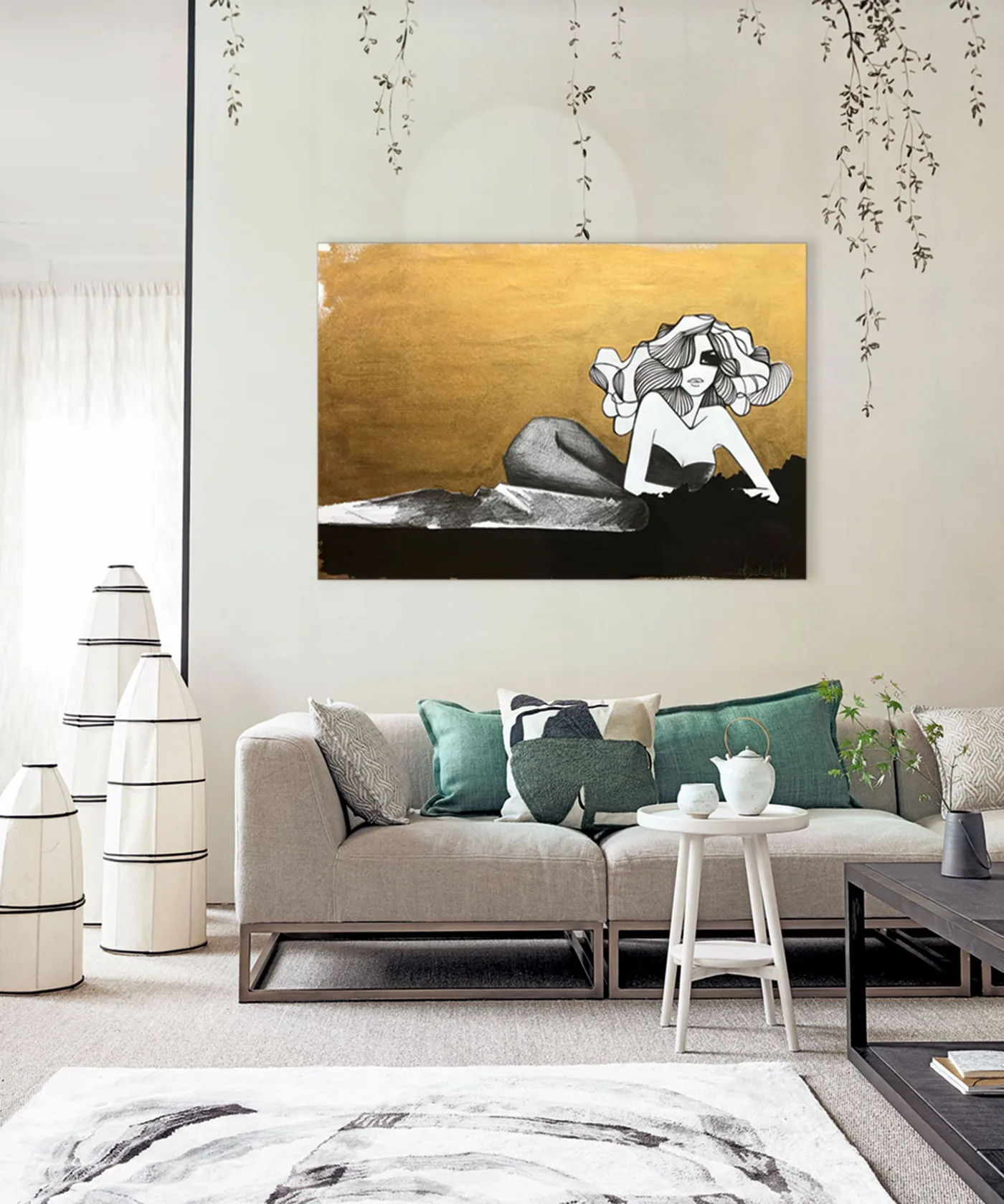 Neutral sofa with green cushions below gold-toned artwork beside pale sculptural floor lights and a dark wood table