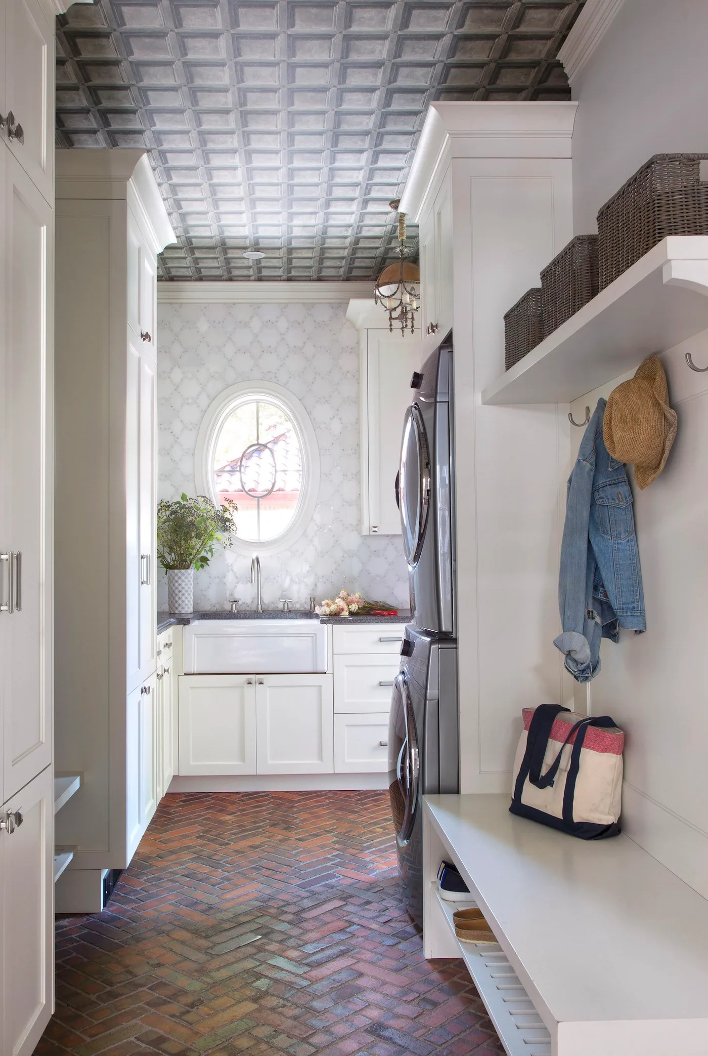 White laundry and mudroom with coat hooks, a bench, warm herringbone flooring and a patterned ceiling.