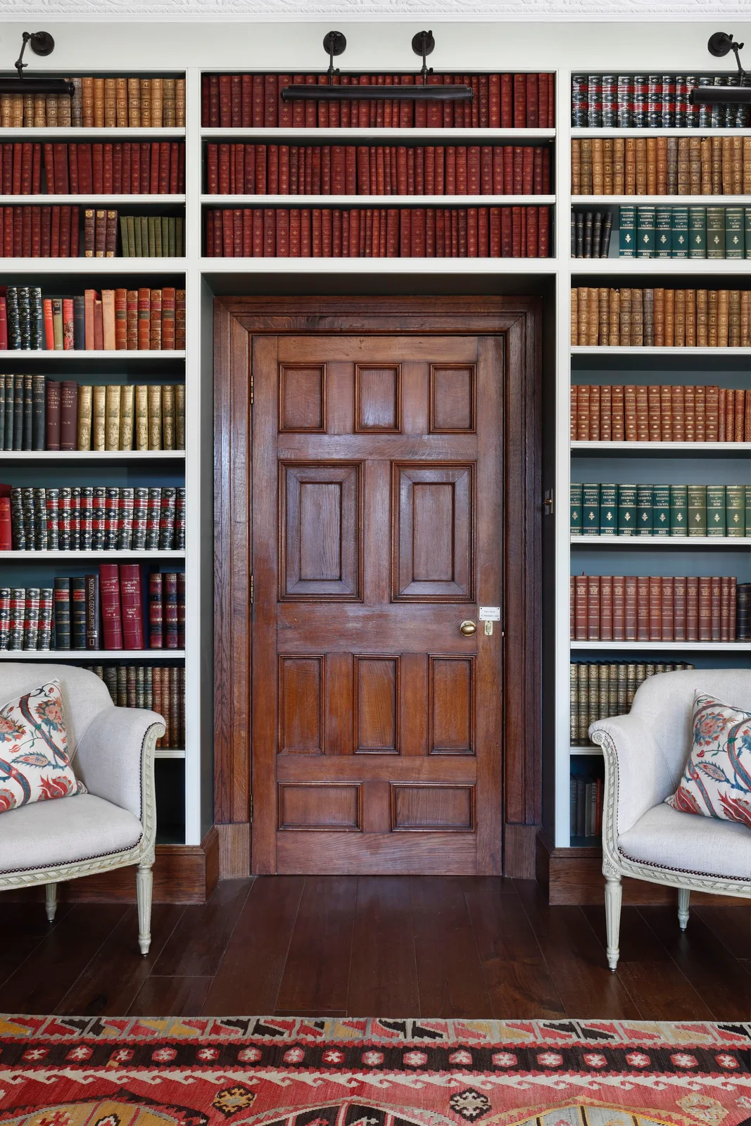 Pale shelves filled with red, brown and green book rows frame a wooden paneled door between two pale chairs