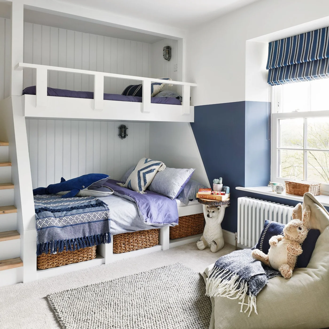 White built-in-style bunk beds have blue bedding and woven baskets below beside navy lower walls, a pale rug and a nearby window