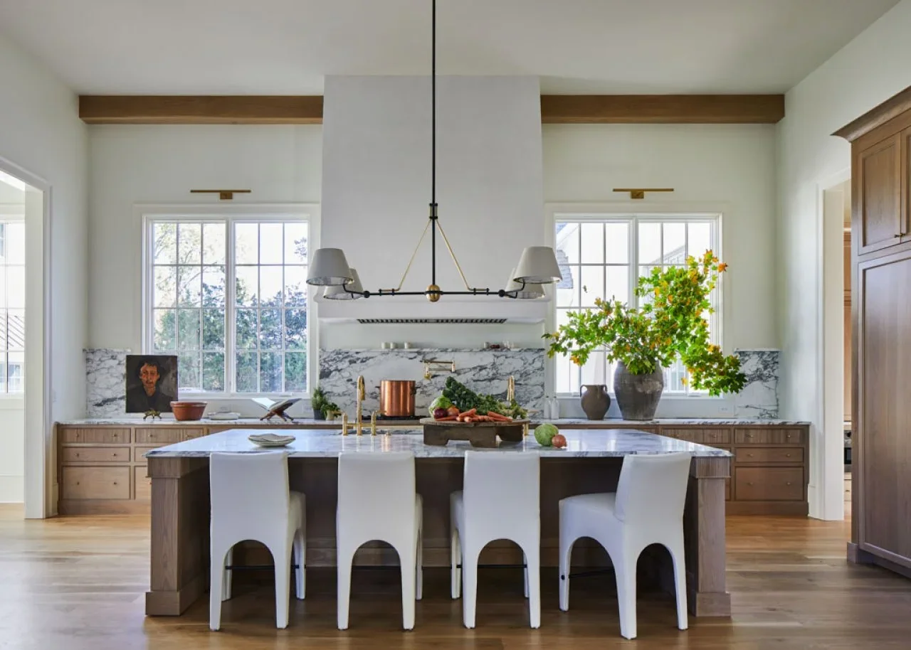 Oak lower cabinets and an island sit beneath a white hood between tall windows, with pale counters and four sculptural white chairs.