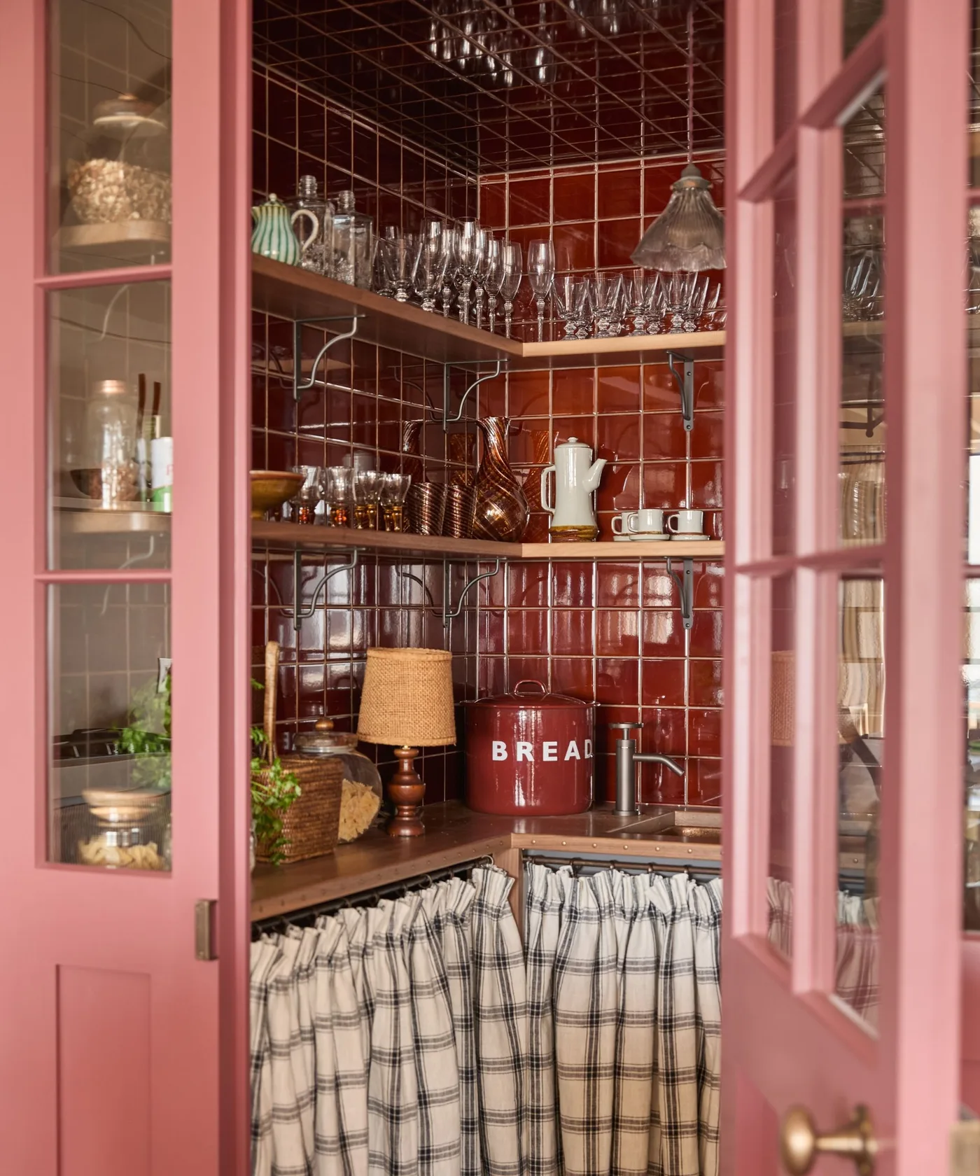 Small country pantry with deep red tile, warm wood shelves, glassware and checked curtain below counter