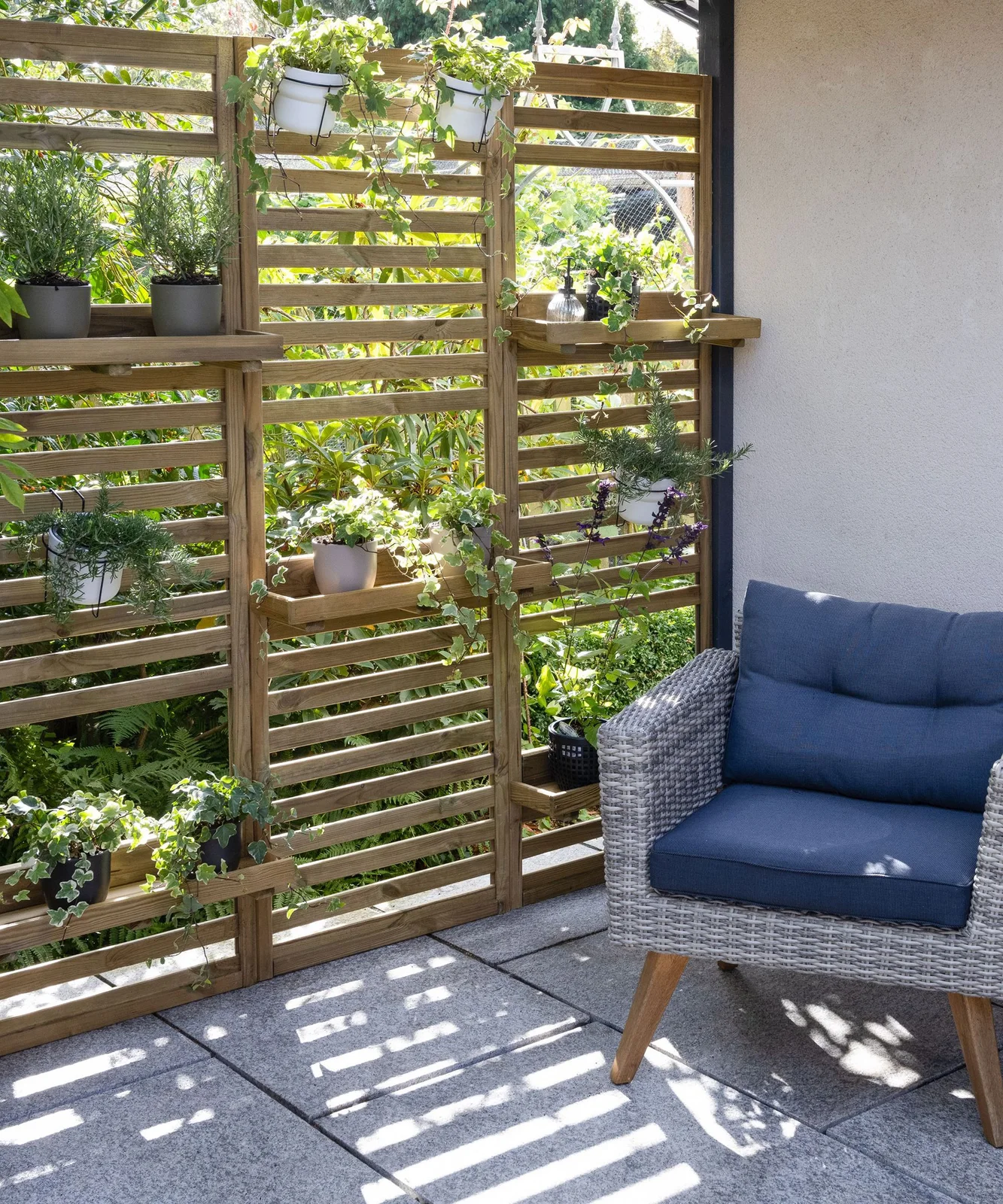 A timber slatted screen with shelves and small pots stands beside a blue-cushioned outdoor chair