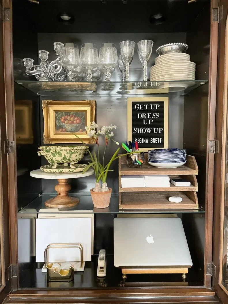Dark dining cabinet containing glassware, plates, a closed laptop and small office supplies