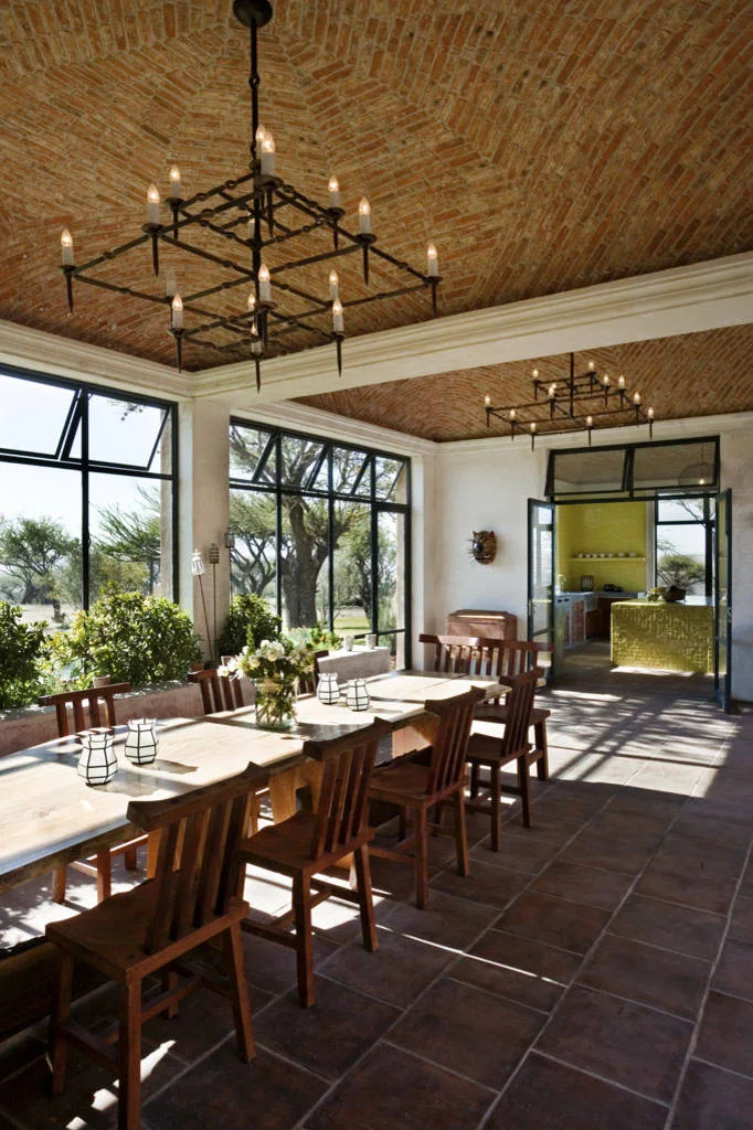 Long wood dining table and matching chairs below dark chandeliers and a patterned brick ceiling beside tall windows.
