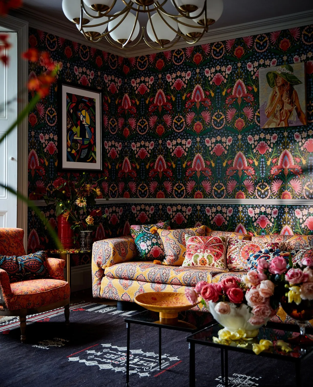 Living room with dark multicolored floral wallpaper, patterned pale sofa, orange armchair, curved overhead light and small tables.