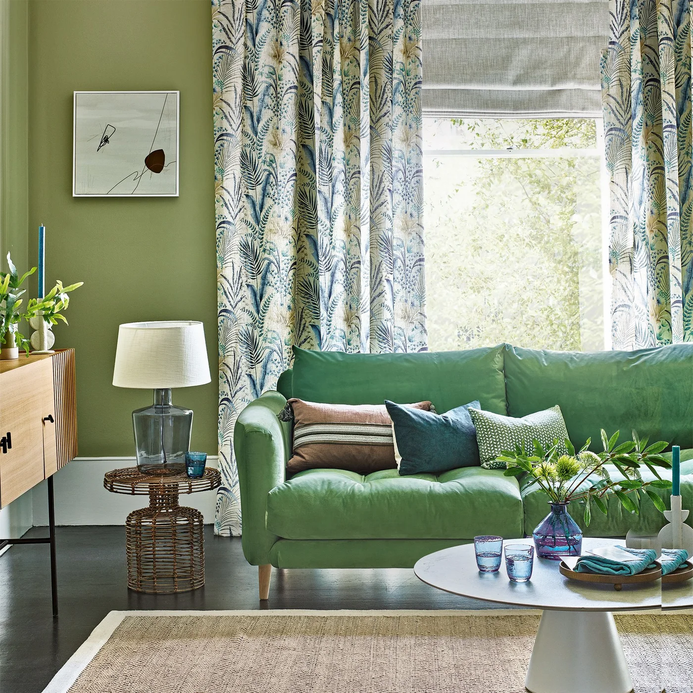 A pale green sofa sits near leafy curtains beside a woven side table and a pale oval coffee table