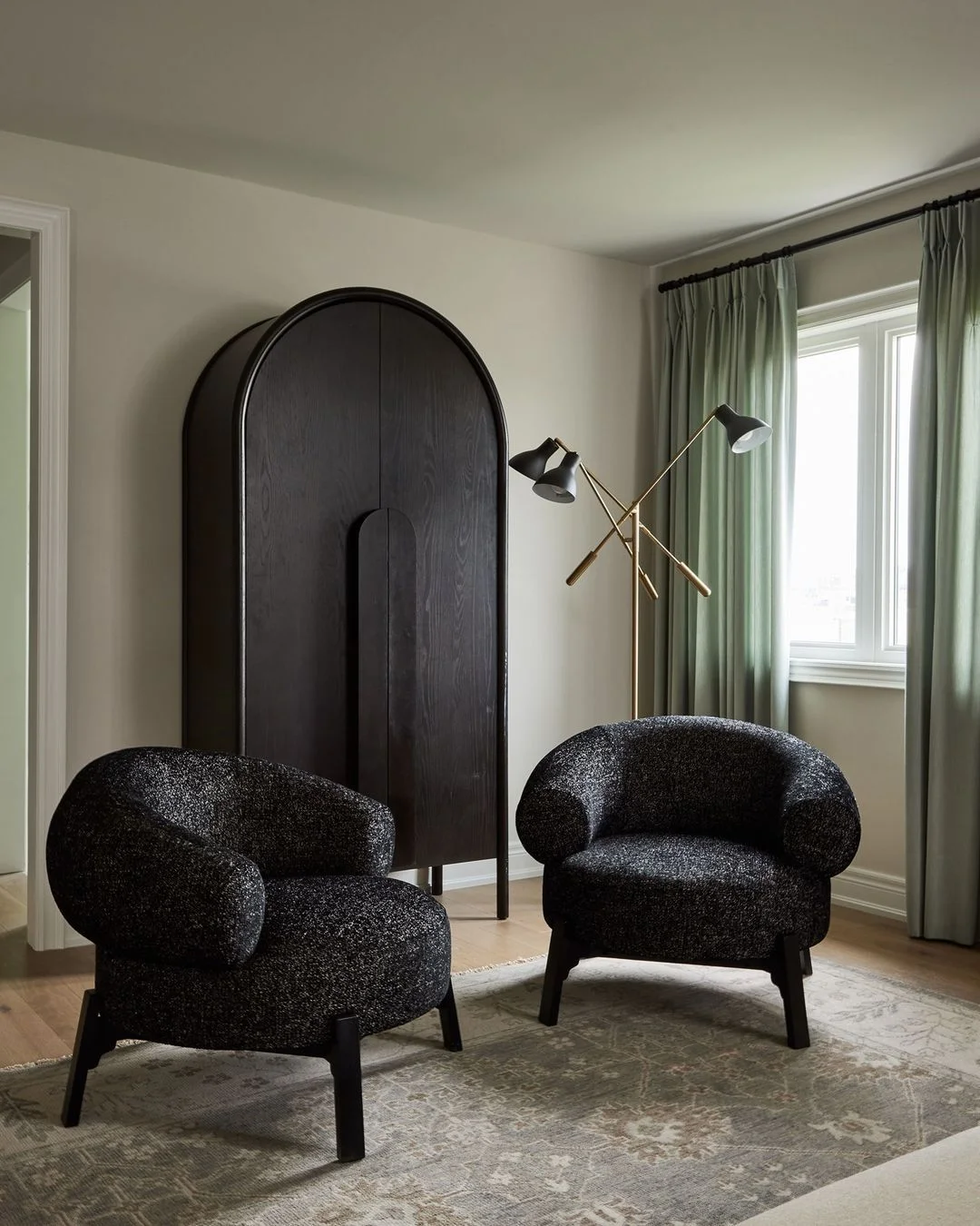 Pale sage curtains hang behind two dark rounded chairs, a tall arched cupboard and a branching floor lamp.