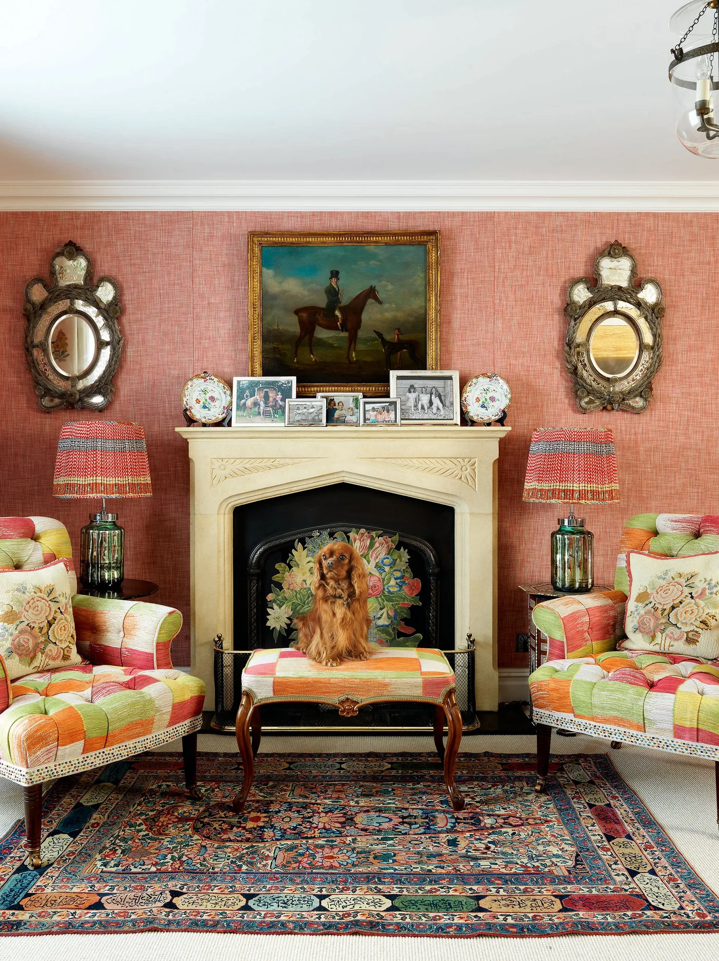 Living room with coral wall, pale fireplace surround, framed photographs, paired ornate mirrors, mixed patterned armchairs, lamps and richly patterned rug.