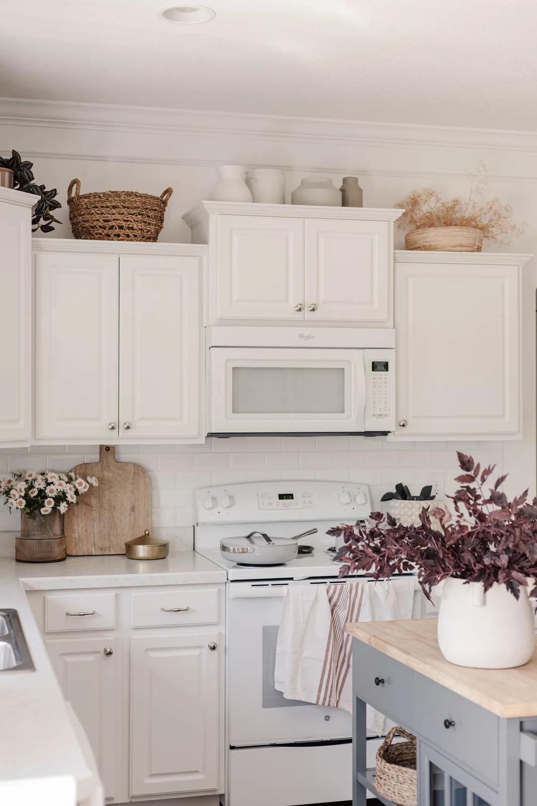 Pale vessels above a raised white cabinet flanked by a woven basket and a shallow basket of dried stems on lower cabinet tops.