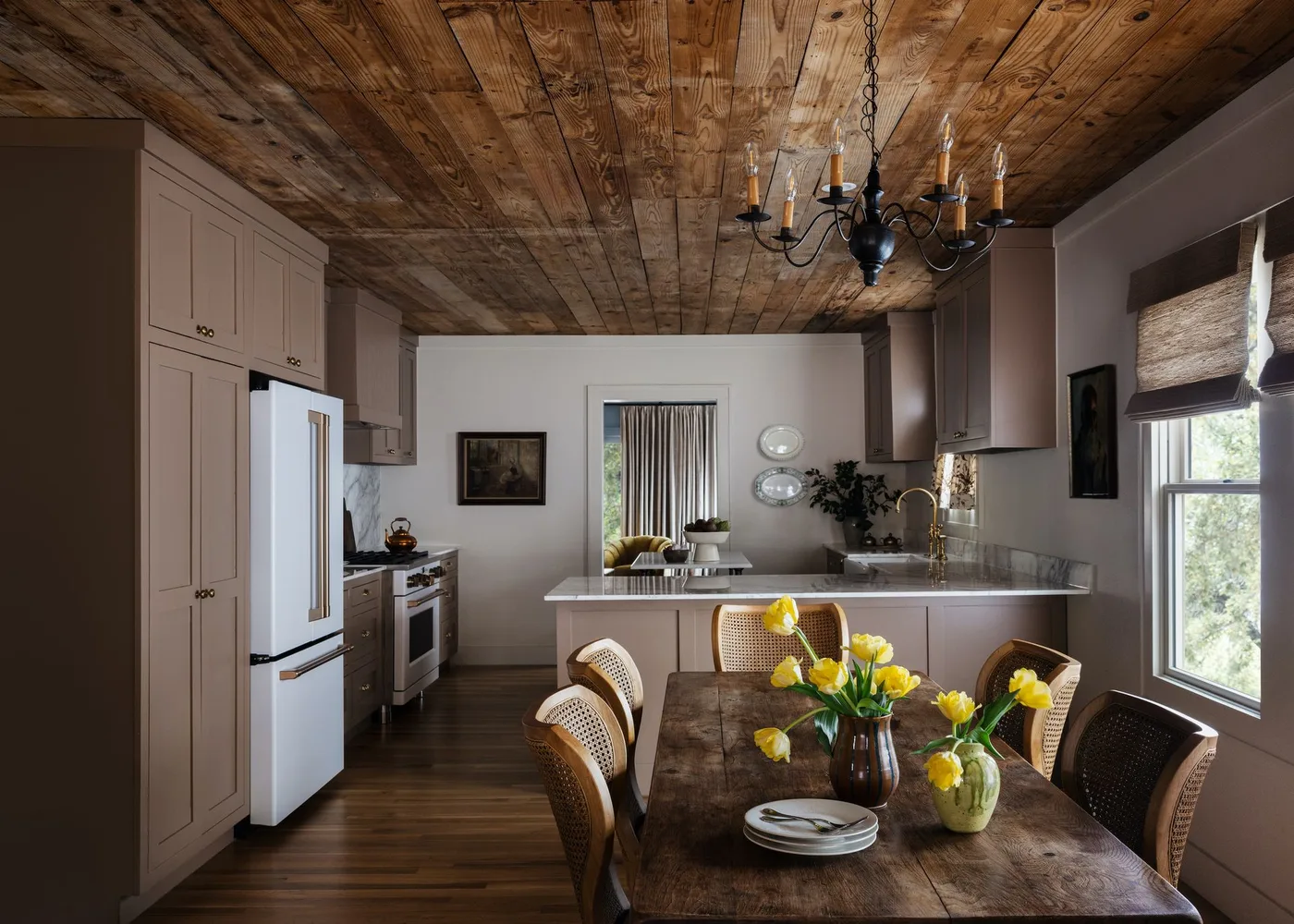 Soft brown kitchen cabinets around a pale island, with a wood ceiling and a dining table with woven chairs.