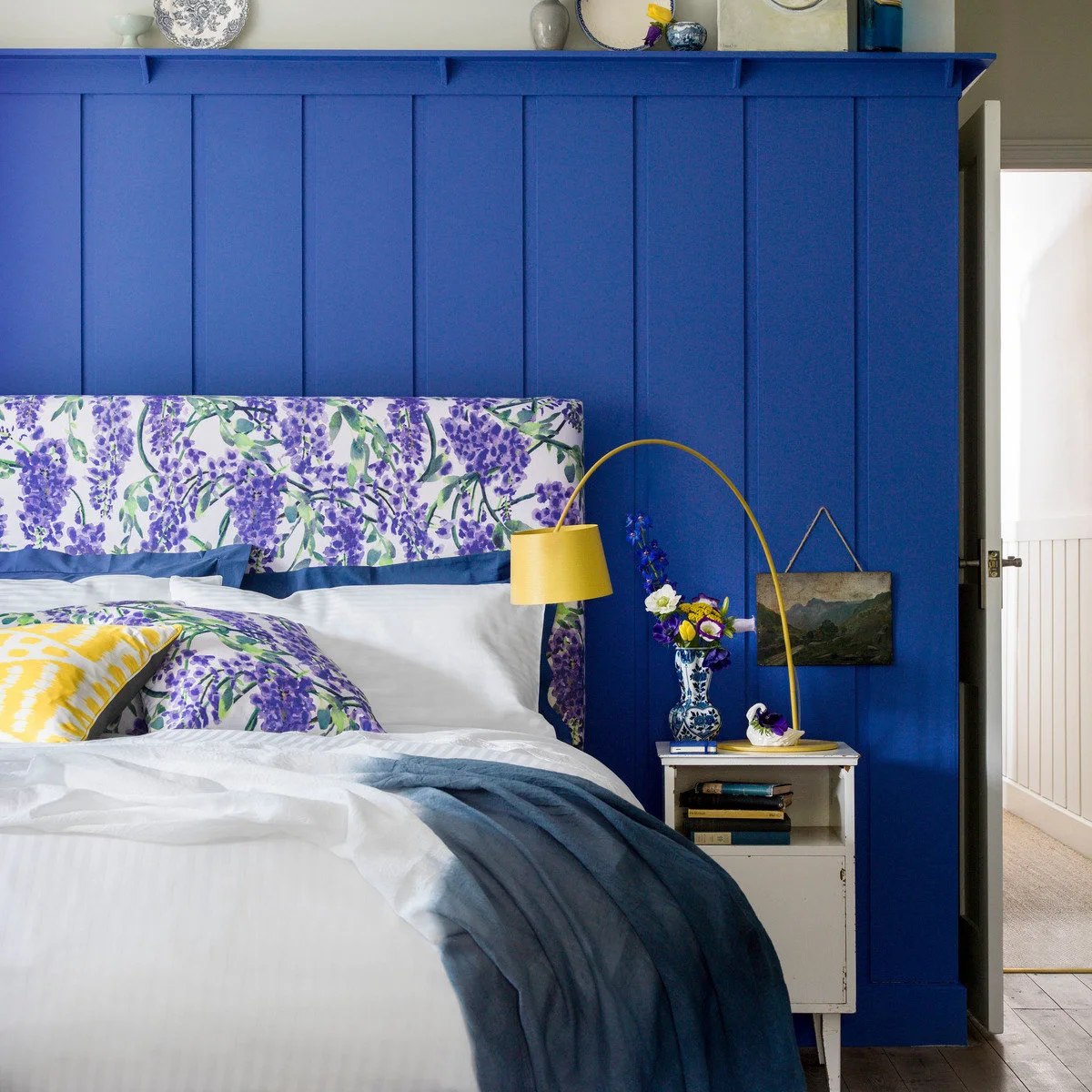 Blue paneled wall behind a floral upholstered headboard with a yellow bedside lamp and white bedding.
