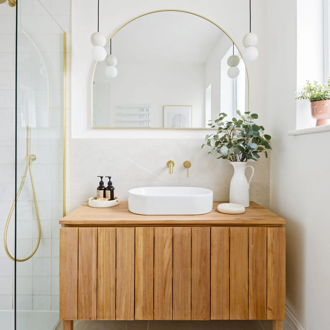 Wood vanity with vertical slat-style fronts below a white vessel basin and large arched mirror.
