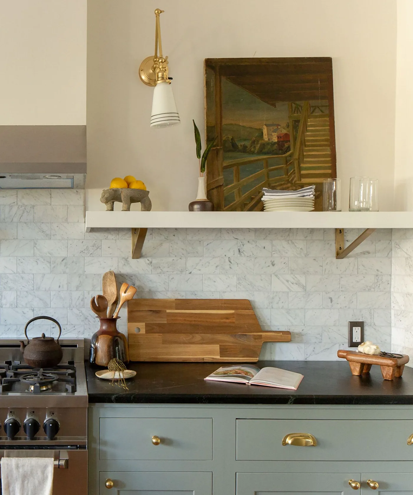 Kitchen with blue-gray lower cabinets, dark counter, pale veined backsplash, open shelf and framed picture.