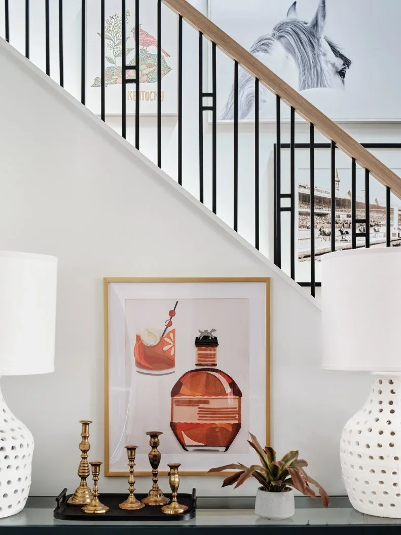 Gold-colored candleholders and a small plant sit on a tray between two pale lamps above a dark sideboard below stair artwork