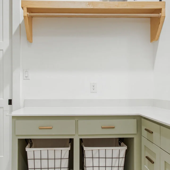 A pale sage counter has two open bays for laundry baskets beneath a wood wall rack and shelf against white walls.