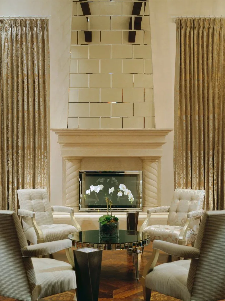 Mirrored rectangular wall tiles above pale carved fireplace surround with gold curtains and white chairs