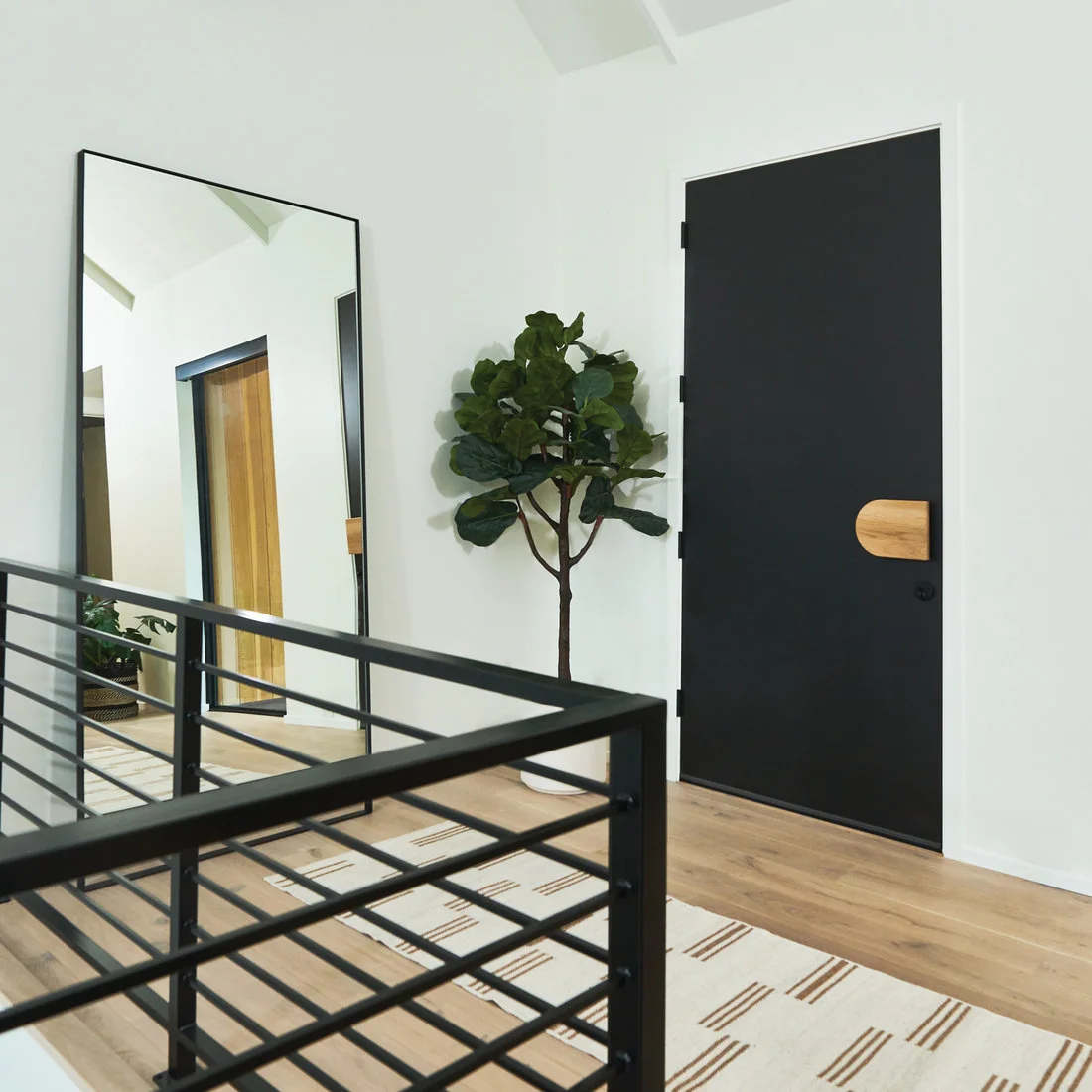 A black entry door with a wood handle faces a tall black-framed mirror beside a plant, pale floor and simple patterned runner near a stair rail.