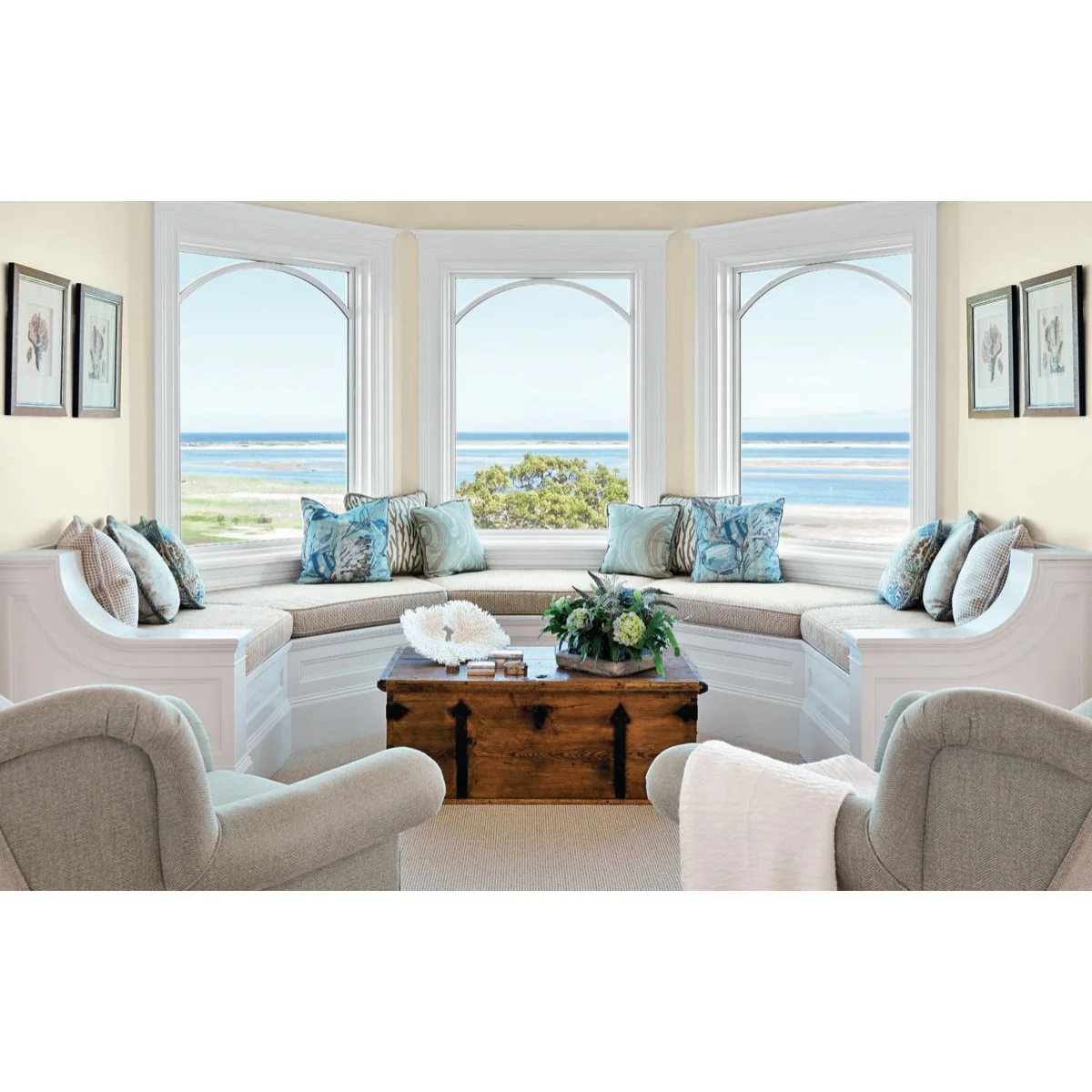 A pale wraparound window seat with blue cushions and a wooden trunk in front of three arched-top windows.