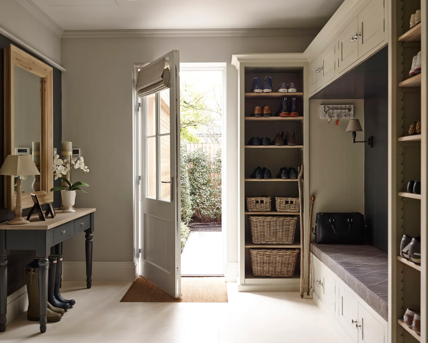 Pale mudroom cabinets surround a cushioned bench with overhead doors, tall shoe shelves and woven baskets beside an exterior door.