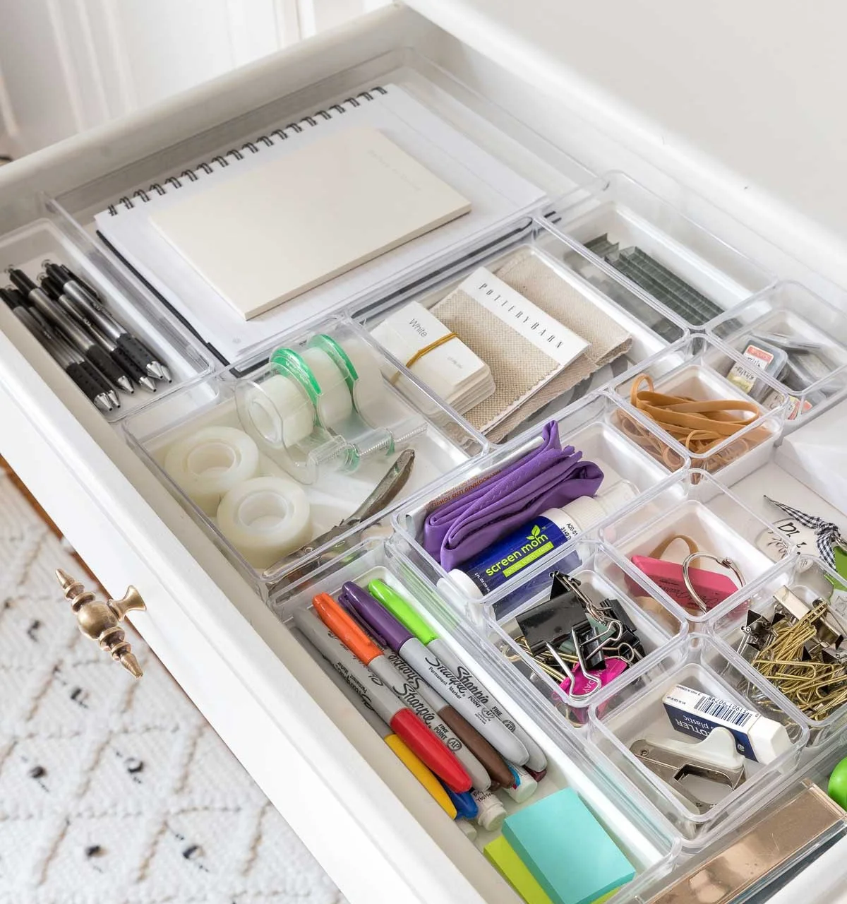 White drawer with clear bins holding a notebook, tape, elastic bands, pens, clips and small accessories
