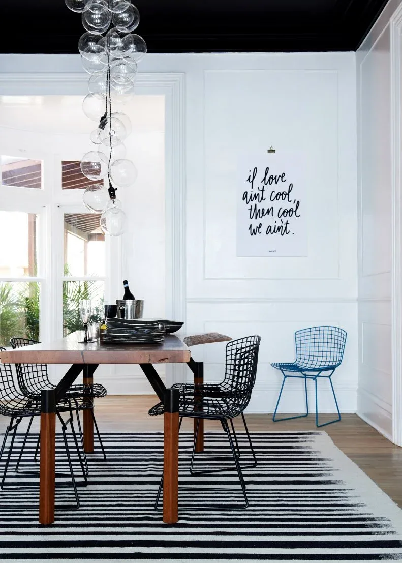 Black-lettered poster on a white paneled dining-room wall, with a wooden table, black wire chairs, one blue chair, striped rug, and glass globe light.