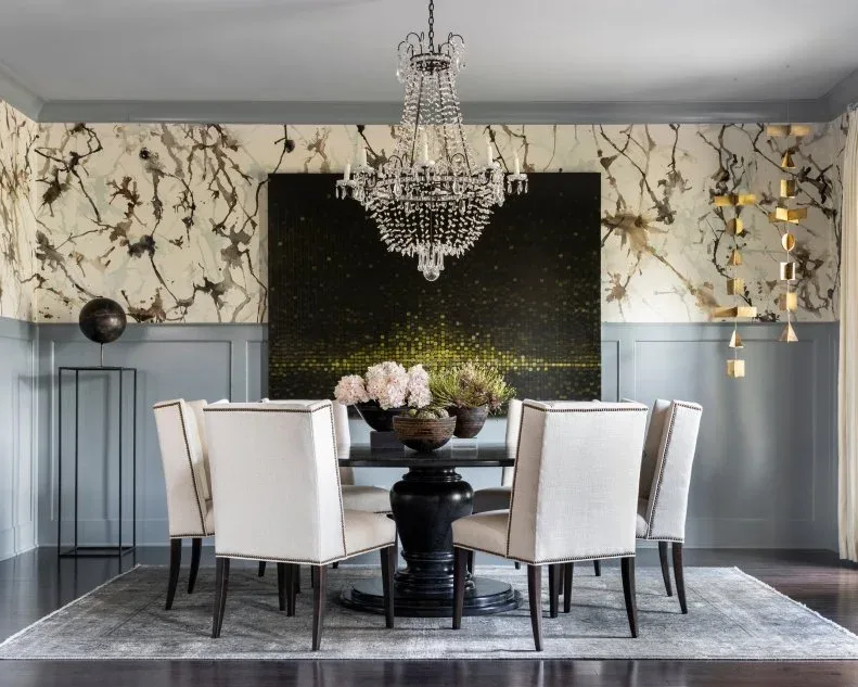 Dining room with gray lower paneling, branch-pattern wallpaper, dark artwork, a round pedestal table and tall pale upholstered chairs