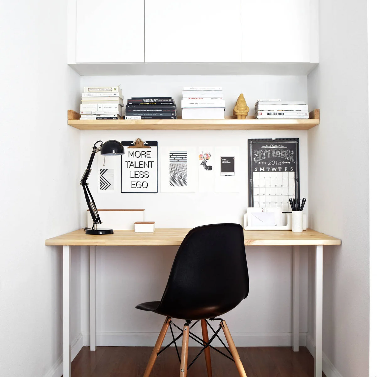 Slim wood-top desk with white legs, a black chair, wood shelf and white wall cabinets