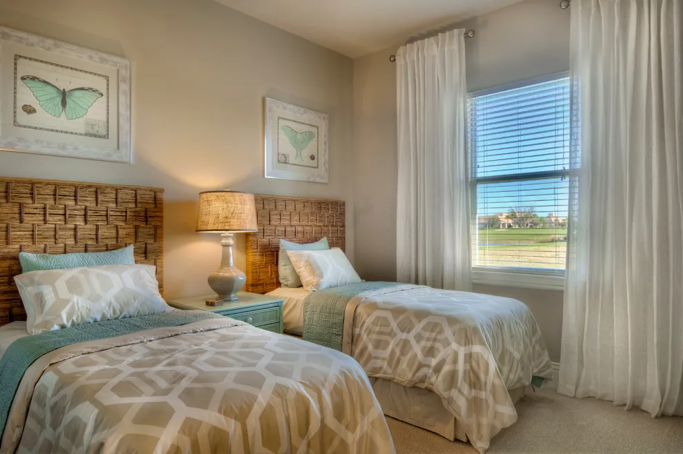 Two beds with beige patterned covers and green-blue accents sit beside pale bedroom walls and curtains