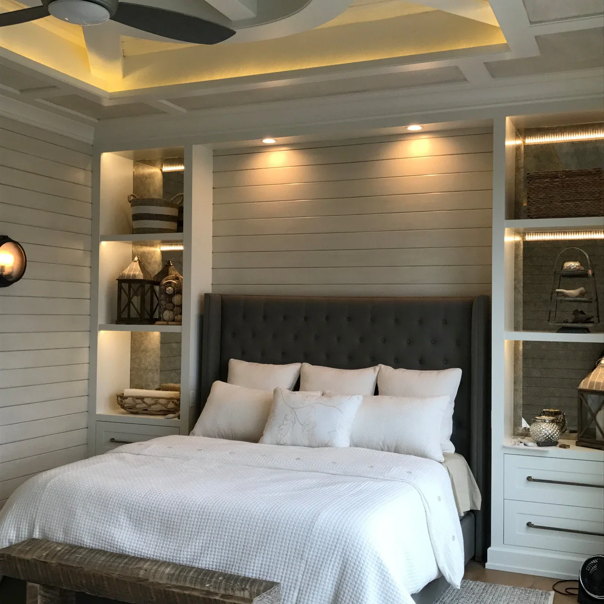 Dark grey tufted headboard between pale built-in shelves and drawers with warm shelf lighting