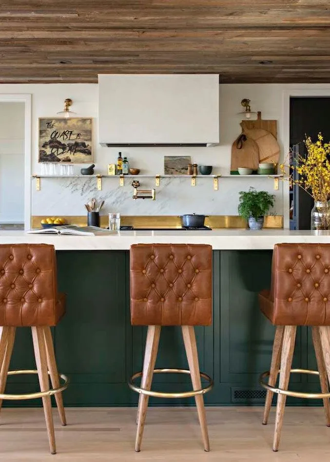 Dark green island with brown tufted stools beneath wood ceiling, facing a white hood and pale veined backsplash